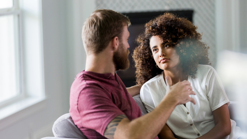 mid adult husband and wife sit on couch talking intently